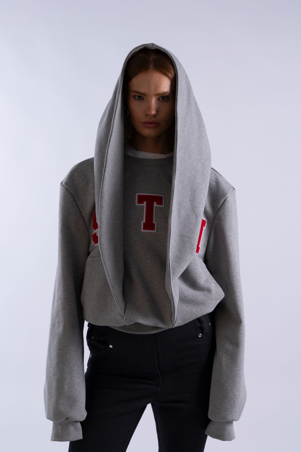 Model wearing the grey STN Collegiate Hoodie by Stadion, featuring oversized structural hood and red collegiate lettering.