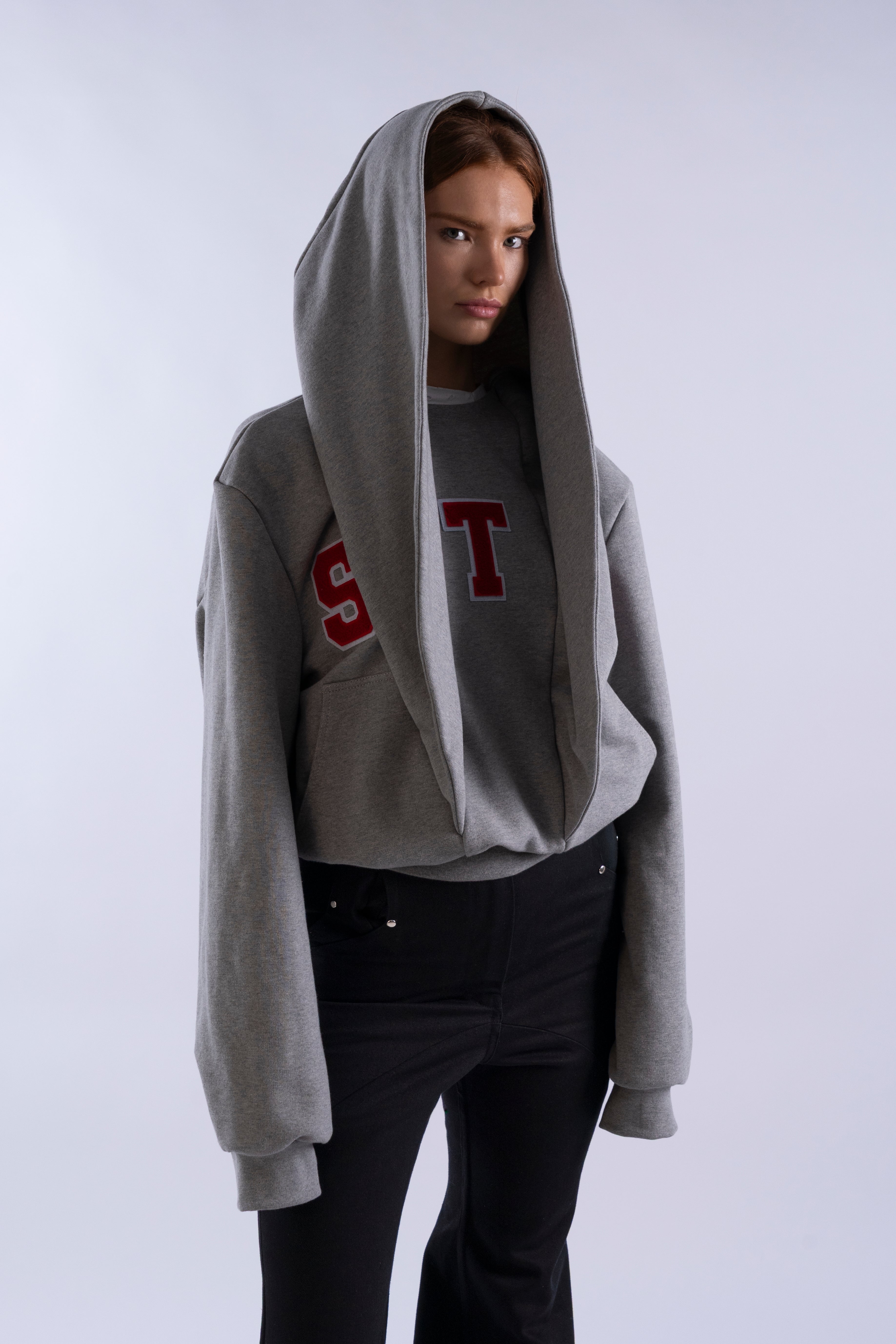 Side angle of model wearing grey STN Collegiate Hoodie by Stadion, featuring oversized hood and red collegiate S T lettering.”